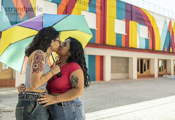 Romantic lesbian couple kissing under multi colored umbrella in front of building