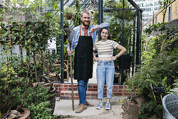 Smiling man in apron standing with hand on head of girlfriend outside greenhouse