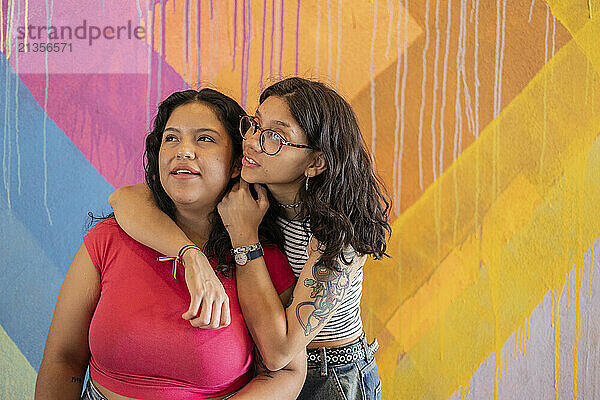 Young woman in eyeglasses standing with arm around girlfriend near painted wall