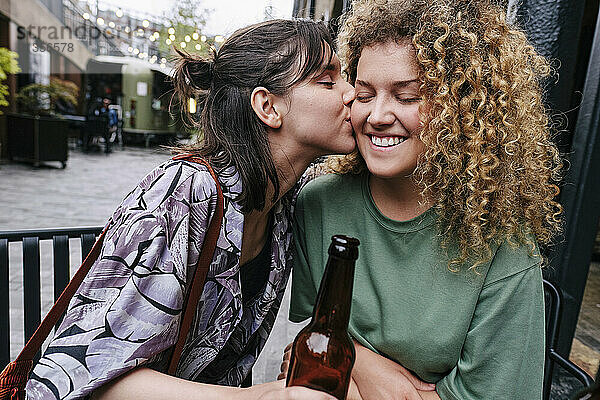 Young woman holding beer bottle and kissing girlfriend at sidewalk cafe