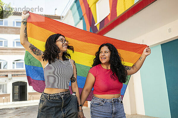 Happy gay couple holding pride flag and walking near building