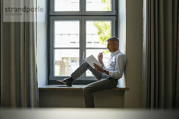 Senior businessman using tablet PC and sitting on window sill in office