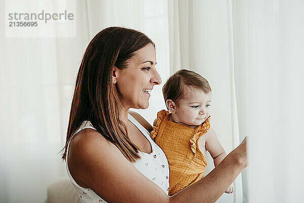 Smiling mother with cute baby girl near window in bedroom