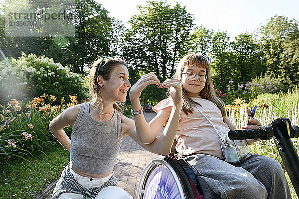 Smiling mother making heart shape with daughter sitting on wheelchair in park