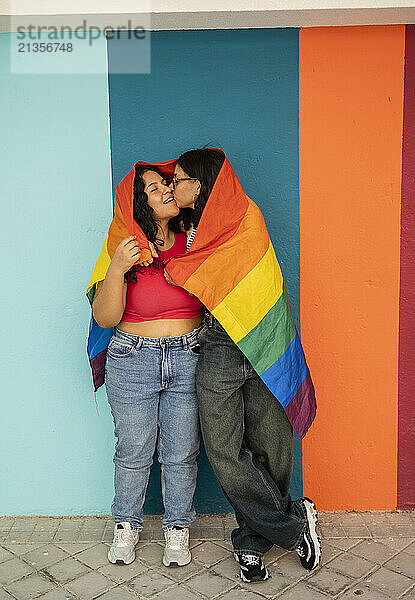 Young woman kissing girlfriend wrapped in pride flag near striped wall