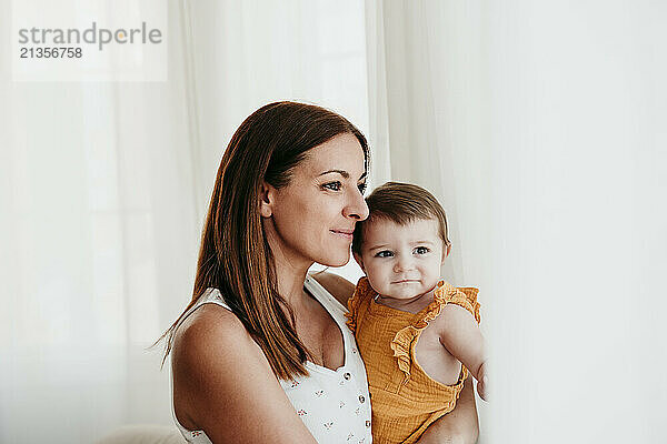 Smiling mother with cute baby girl near window in bedroom at home