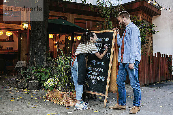 Young woman writing on chalkboard with co-owner standing outside coffee shop