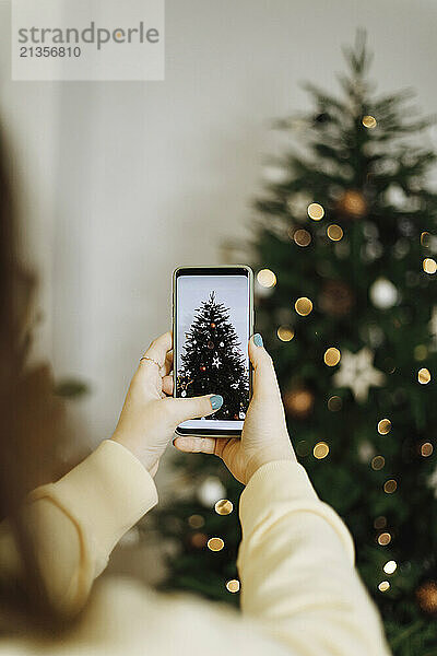 Young woman photographing Christmas tree through smart phone at home