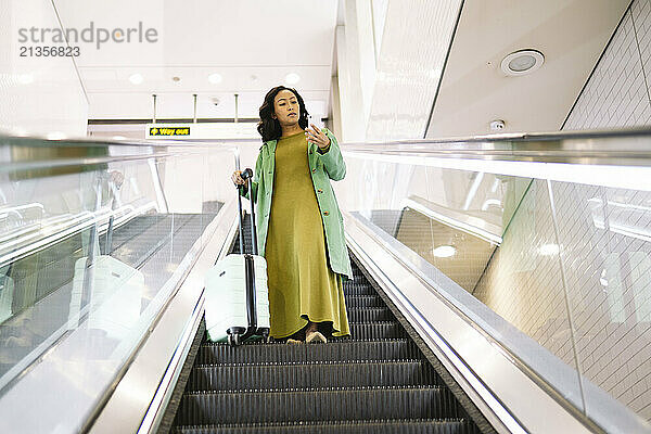 Woman standing with suitcase and using mobile phone on escalator