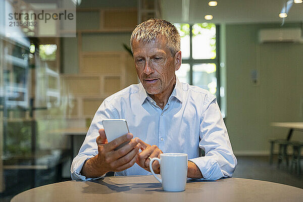 Smiling senior businessman using smart phone at table in office cafeteria