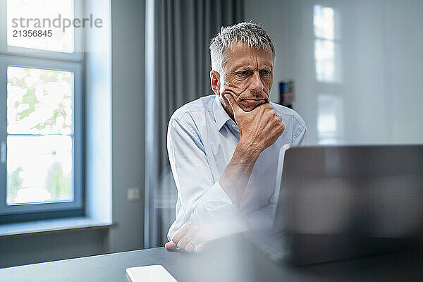 Senior businessman sitting with hand on chin and using laptop at desk