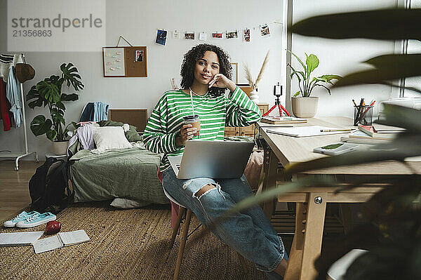 Smiling teenage girl sitting with laptop and hot chocolate at home