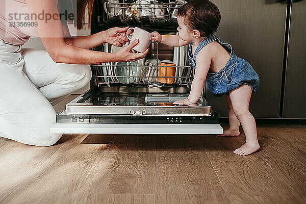 Baby girl helping mother in unloading utensils from dishwasher