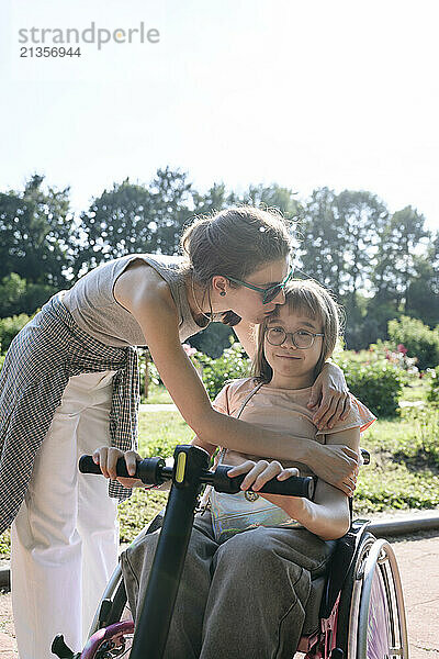 Mother kissing daughter sitting on wheelchair at park