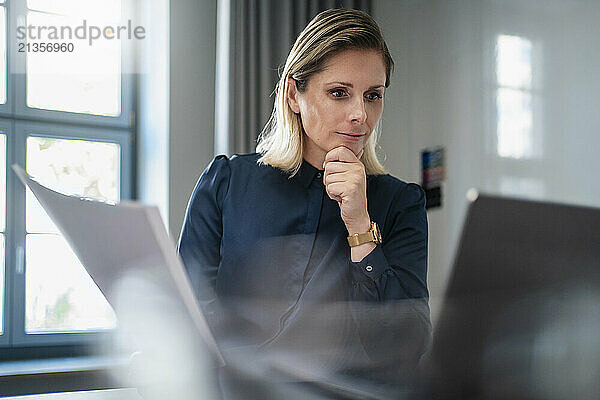 Businesswoman holding document and working on laptop at desk