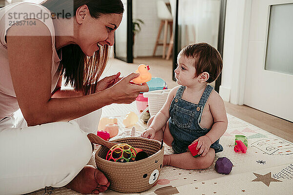 Happy mother holding toy duck and playing with daughter sitting on rug at home