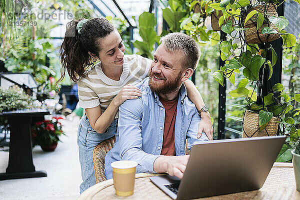 Woman with arm around boyfriend sitting near laptop in greenhouse