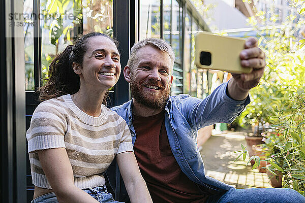 Happy boyfriend taking selfie with girlfriend near greenhouse