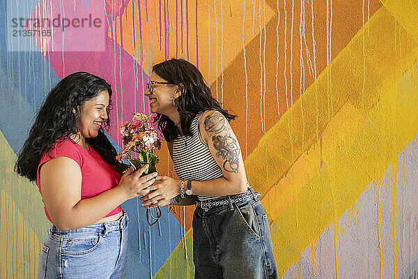 Happy woman giving bouquet of flowers to girlfriend standing near painted wall