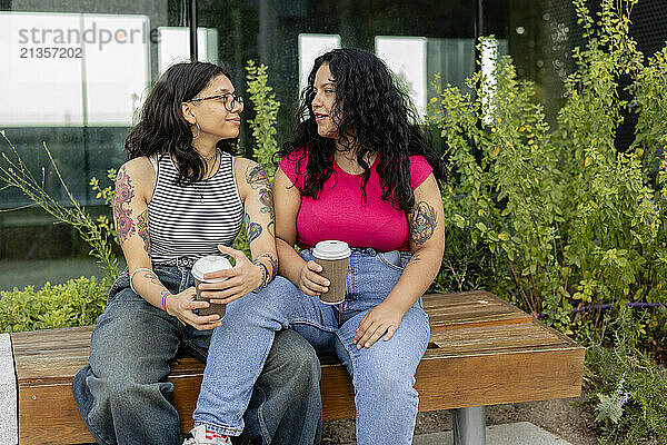 Young gay couple holding disposable cups talking and sitting on table