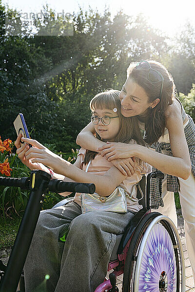 Mother embracing daughter sitting on wheelchair and taking selfie at sunny day