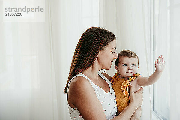 Affectionate mother with cute baby girl near window in bedroom at home