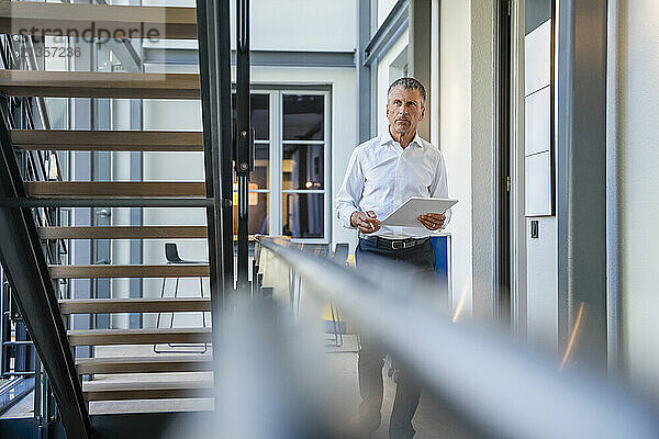 Senior businessman walking with tablet PC near staircase in office corridor