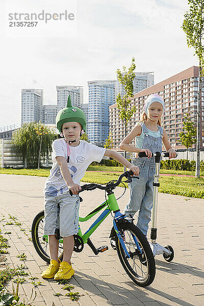 Siblings with bicycle and push scooter standing on footpath