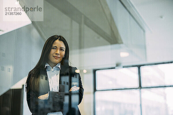 Confident businesswoman with arms crossed seen through glass at workplace