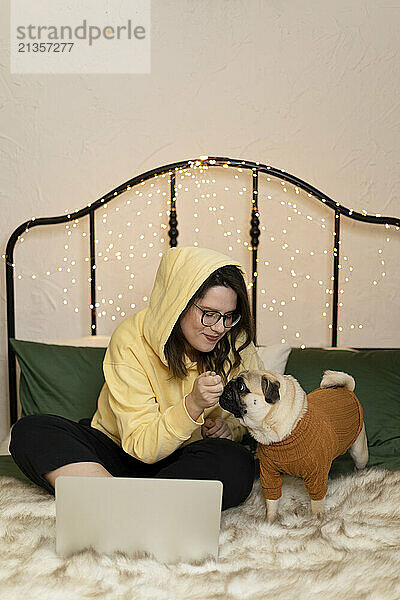 Young woman sitting with laptop and giving treat to pug dog on bed at home