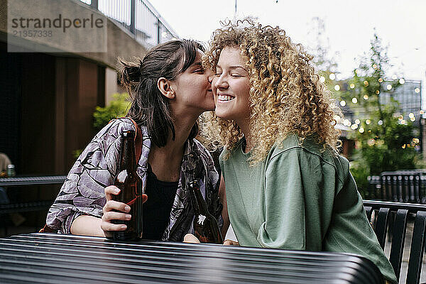 Young woman holding beer bottle and kissing girlfriend at table