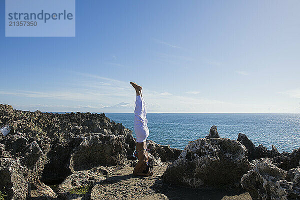 Yoga teacher doing headstand on cliff of Mt Agung under blue sky