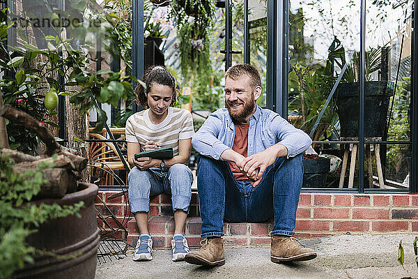 Smiling man sitting with girlfriend using tablet PC outside greenhouse