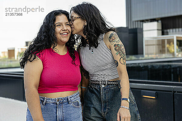 Young woman with tattooed arm kissing girlfriend standing on bridge