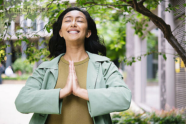 Happy woman standing with eyes closed and hands clasped near tree