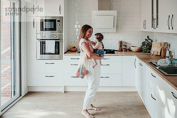 Mother standing with mesh bag and carrying daughter in kitchen at home