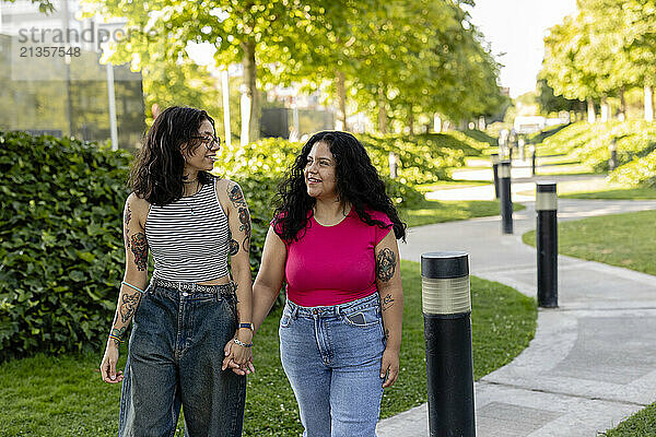 Smiling lesbian couple holding hands and walking on footpath in garden