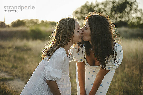 Mother kissing daughter on field at sunset