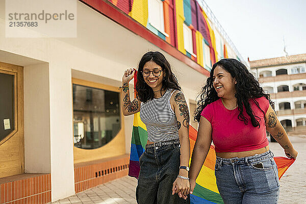 Cheerful gay couple holding hands and enjoying with pride flag near building