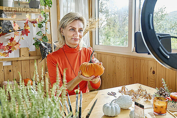 Blond mature woman showing DIY pumpkin and vlogging through smart phone at home