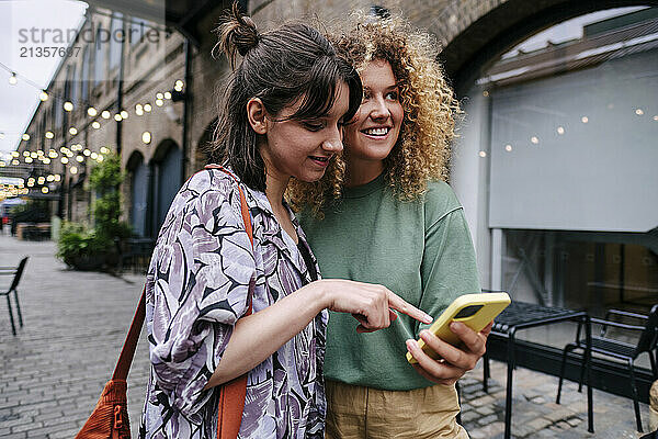 Smiling woman using smart phone with friend standing near building