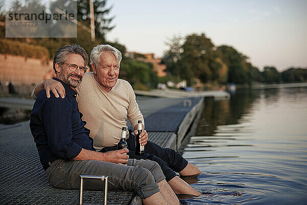 Smiling senior friends sitting with beer bottles on jetty