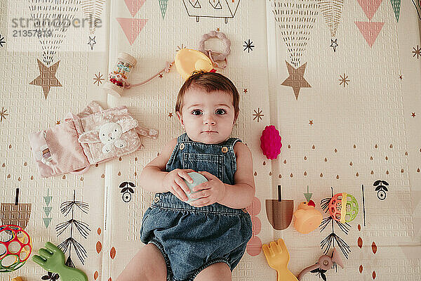 Cute baby girl in bib overalls lying on mat with toys at home