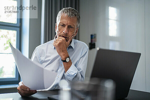 Senior businessman reading documents near laptop at desk in office