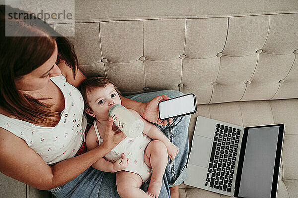 Freelancer mother using smart phone and feeding daughter on sofa at home