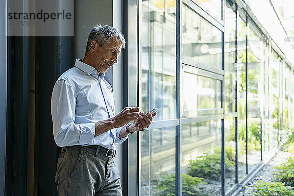 Senior businessman leaning on glass and using smart phone in office