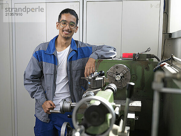 Smiling apprentice wearing jacket leaning on machine at workshop