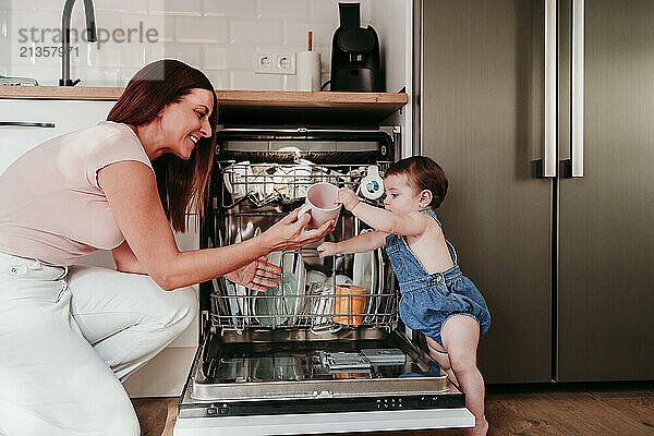 Baby girl helping mother in unloading utensils from dishwasher at home