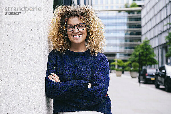 Happy woman leaning with arms crossed on wall in front of buildings