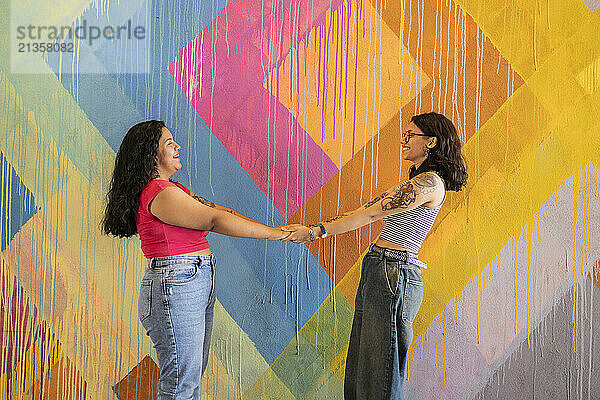 Happy young lesbian couple holding hands and standing in front of painted wall